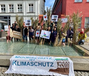 Presse- und Öffentlichkeitsarbeit zum Thema Moor bei der Michael Succow Stiftung (MSF) verantworten, moorbezogene PR-Aktivitäten koordinieren und diese in die Kommunikation des Greifswald Moor Centrum (GMC) integrieren sind wesentliche Aufgaben der PR-Stelle. Hier Mitarbeitende der MSF und des GMC bei einer Aktion im Greifswalder Fischmarktbrunnen. Foto: S. Kagemann/ MSF